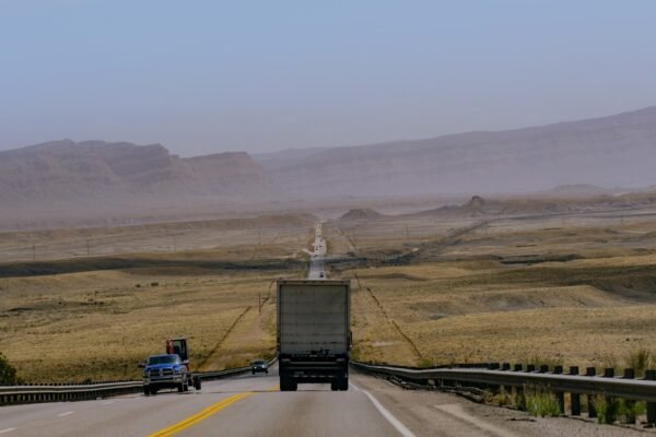 Long desert highway stretching toward distant mountains with a truck and vehicles travelling along it, representative of the Makran coastal road network patrolled by customs enforcement near Gwadar