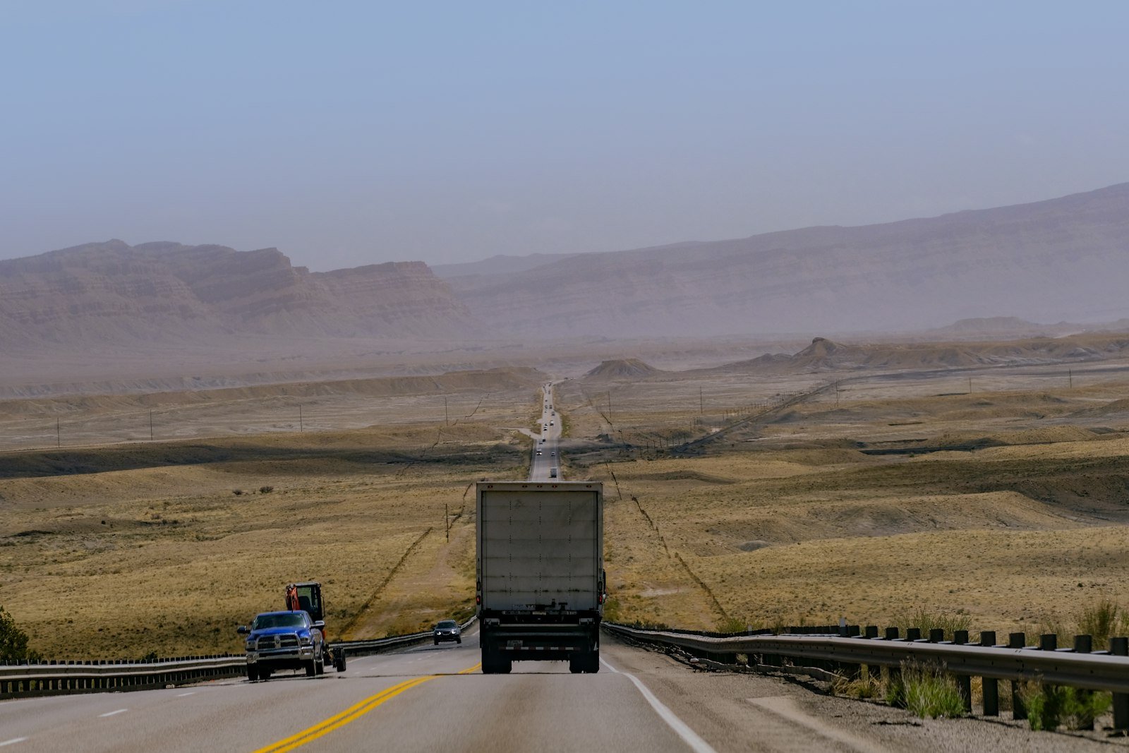 Long desert highway stretching toward distant mountains with a truck and vehicles travelling along it, representative of the Makran coastal road network patrolled by customs enforcement near Gwadar